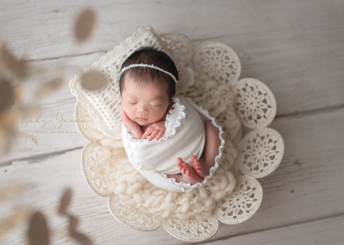 Newborn baby wrapped in white fabric on a floral crochet prop on a wooden floor.