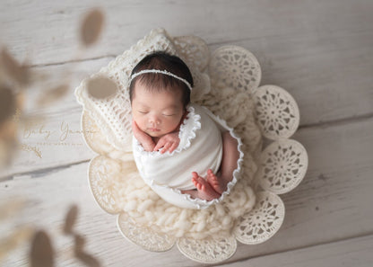 Newborn baby wrapped in white fabric on a floral crochet prop on a wooden floor.