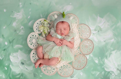 Newborn baby in a green outfit lying on a decorative floral bed against a matching green background