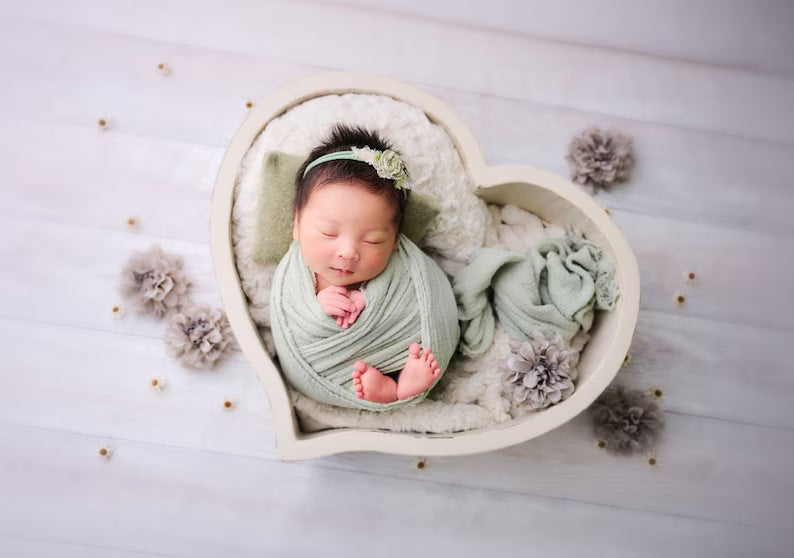 Newborn baby wrapped in a light green blanket, lying in a heart-shaped bowl on a white surface with decorative flowers.